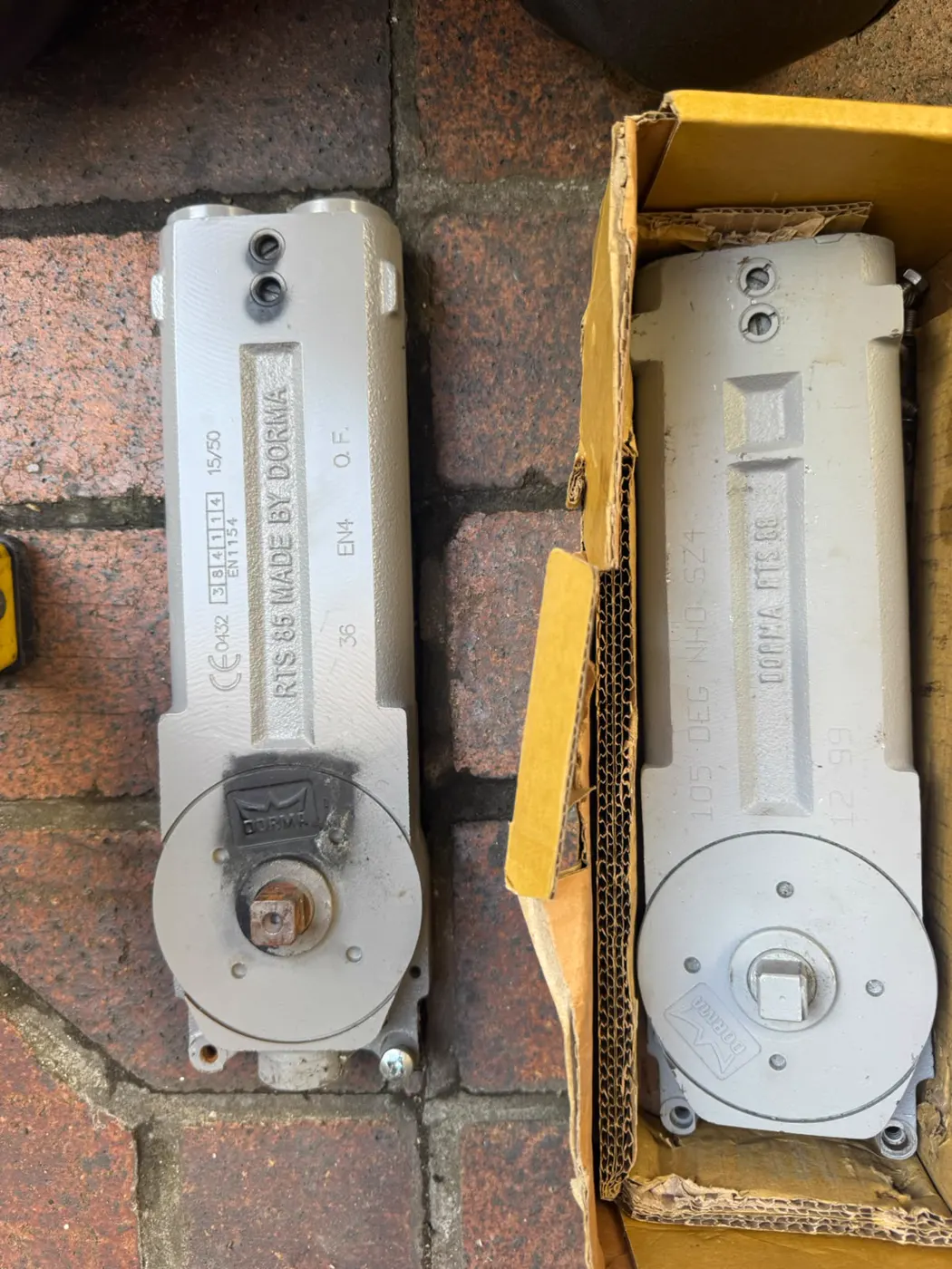 Old corroded Dorma RTS 85 transom closer beside its new boxed replacement on the courtyard paving at the Flux Museum East London